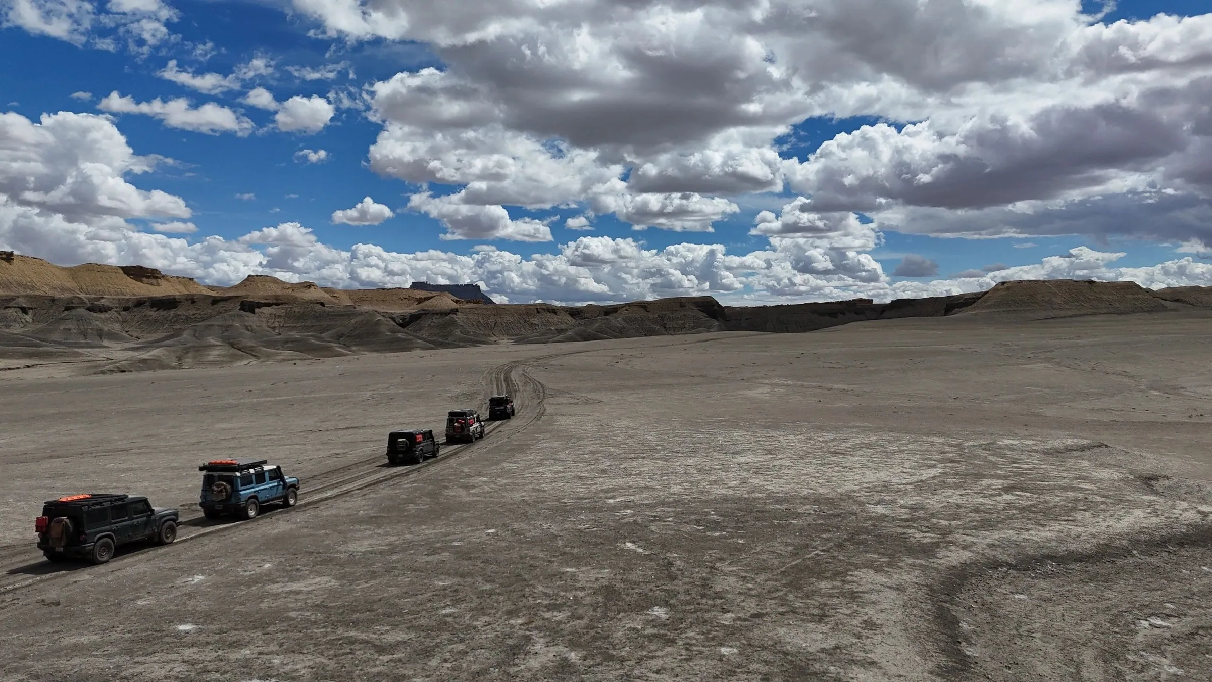 Drone view of a vehicle convoy crossing gray badlands under puffy clouds