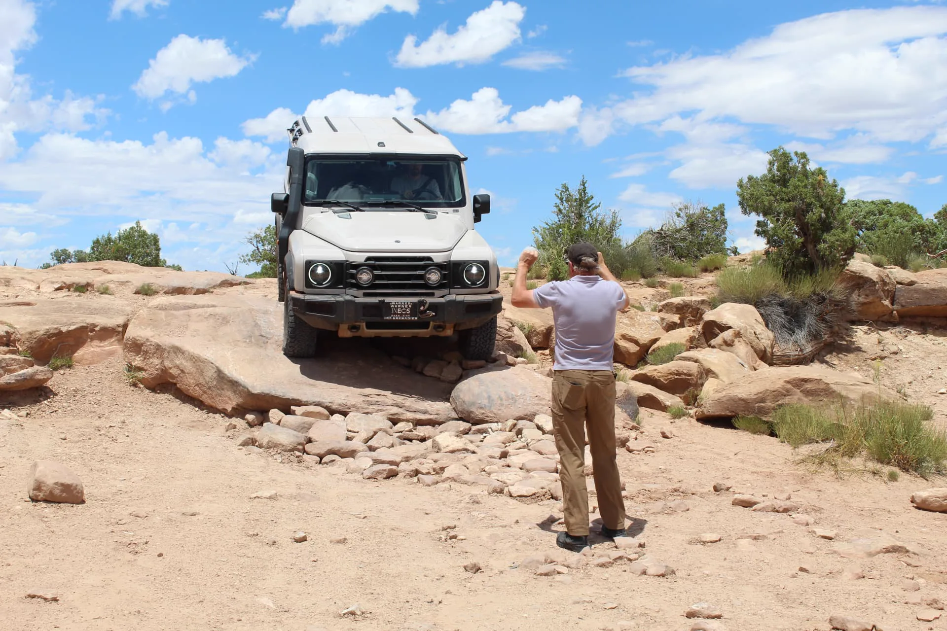 Mike Reed, seen from behind with arms raised, spotting a sand-colored 4x4 as it descends a sandstone rock shelf